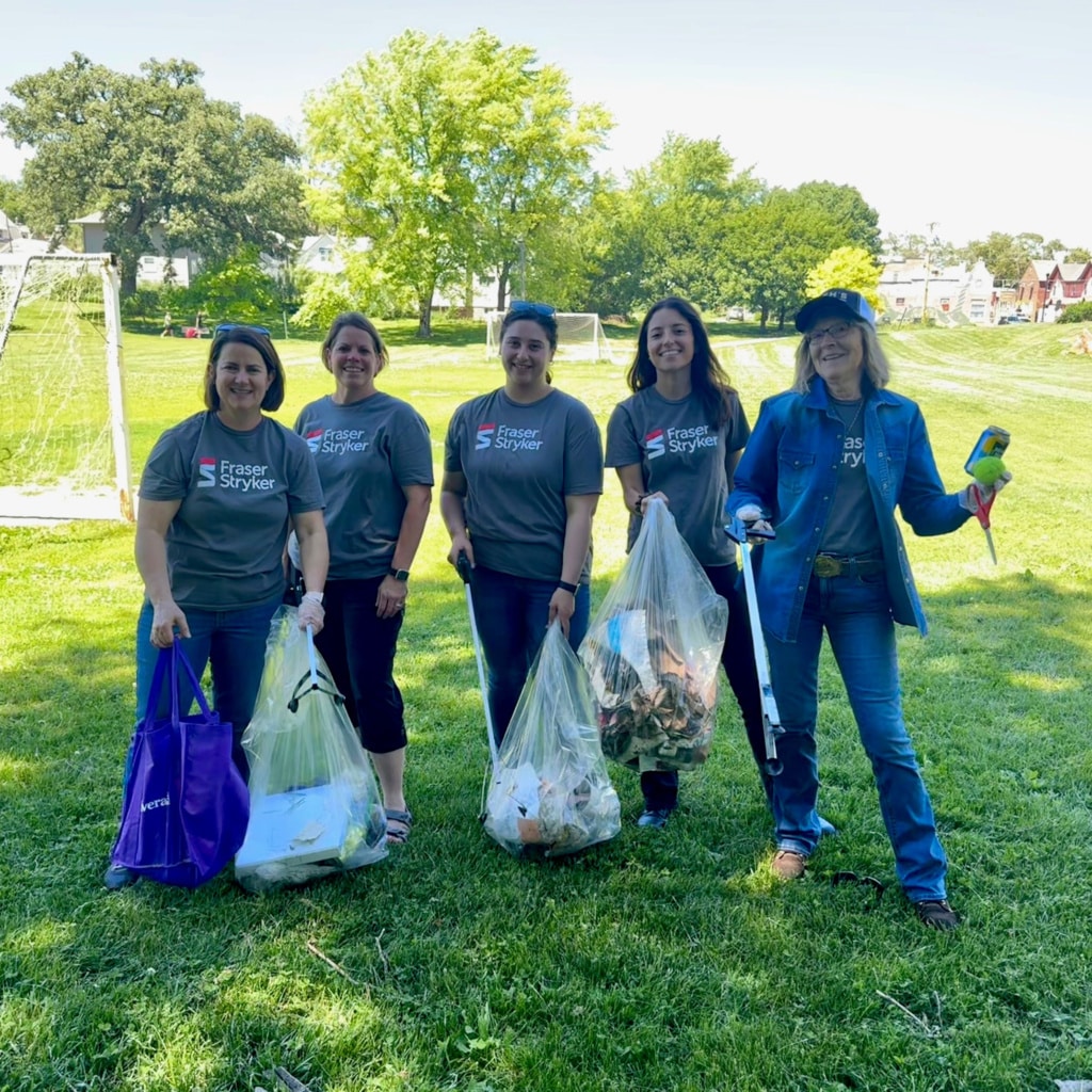 Five Fraser Stryker employees wearing grey Fraser Stryker shirts standing in a park holding bags of trash picked up during a cleanup day for Keep Omaha Beautiful