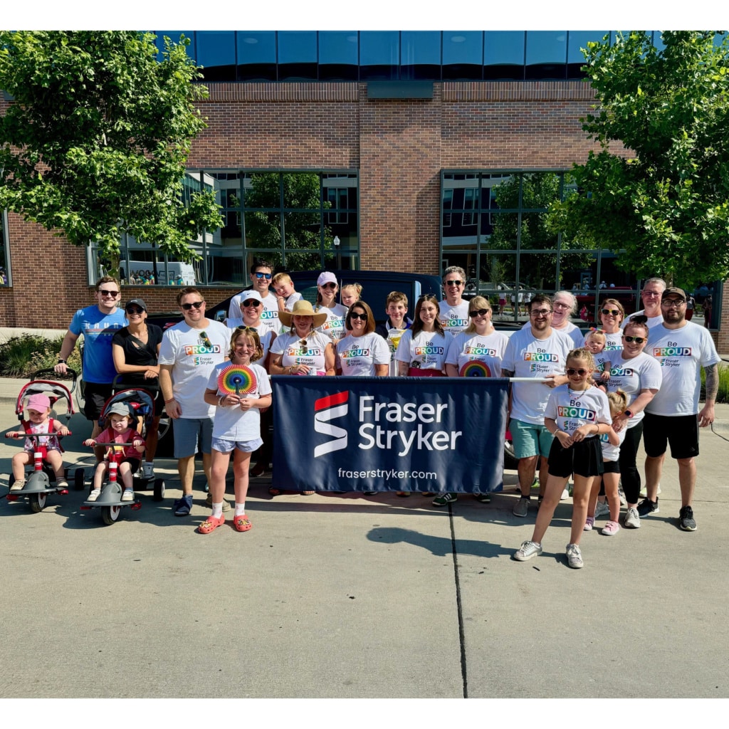 Group of people standing outside holding a Fraser Stryker banner with logo and website. Individuals are wearing white shirts with the Fraser Stryker logo and the words "Be PROUD" written on the front of the shirt.