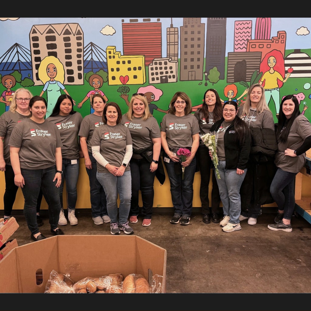 Eleven Fraser Stryker employees wearing grey shirts posing for a photo in front of a city mural painting at Together Omaha